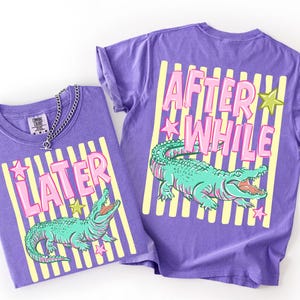May include: Two violet t-shirts with a graphic design. One shirt has the word "LATER" and the other says "AFTER WHILE" in pink letters. Both shirts feature a green alligator illustration and yellow stripes.