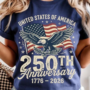 Pode incluir: Uma t-shirt azul-marinho com um gr&aacute;fico de uma &aacute;guia-de-cabe&ccedil;a-branca em frente a uma bandeira americana. O texto diz UNITED STATES OF AMERICA, 250TH Anniversary e 1776 - 2026. O design usa uma fonte de estilo vintage e cores patri&oacute;ticas.