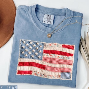 May include: A blue jean colored t-shirt with a sewn-on American flag design. The flag features white stars on a blue background and red and white stripes made of patterned fabric. A gold necklace with a pendant rests on the shirt.