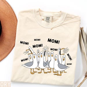 K&ouml;nnte beinhalten: Cremefarbenes T-Shirt mit einer Grafik von Cartoon-M&ouml;wen, die "MOM!" schreien. Die M&ouml;wen sind wei&szlig; mit grauen Fl&uuml;geln und gelben F&uuml;&szlig;en. Der Text "MOM!" ist schwarz, mit dem Comfort Colors-Etikett sichtbar.