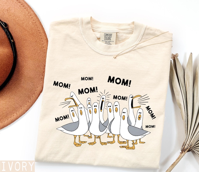 K&ouml;nnte beinhalten: Cremefarbenes T-Shirt mit einer Grafik von Cartoon-M&ouml;wen, die "MOM!" schreien. Die M&ouml;wen sind wei&szlig; mit grauen Fl&uuml;geln und gelben F&uuml;&szlig;en. Der Text "MOM!" ist schwarz, mit dem Comfort Colors-Etikett sichtbar.