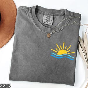 May include: A gray Comfort Colors t-shirt with a yellow sun and blue waves graphic. A silver necklace with a pendant is draped across the shirt. The shirt is folded and displayed on a white surface.
