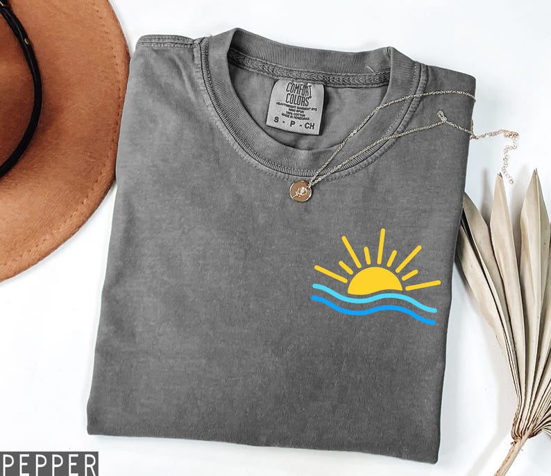May include: A gray Comfort Colors t-shirt with a yellow sun and blue waves graphic. A silver necklace with a pendant is draped across the shirt. The shirt is folded and displayed on a white surface.