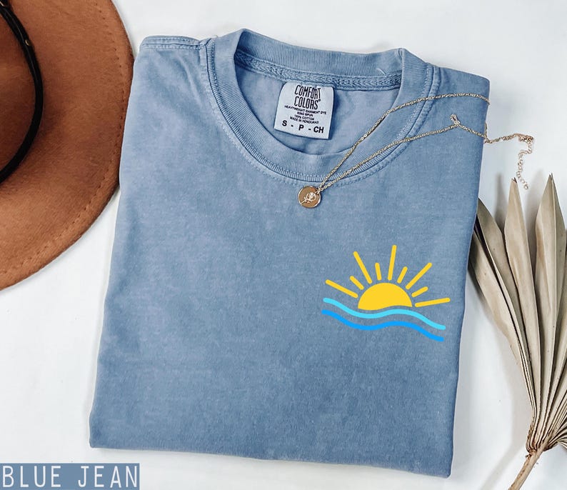 May include: Blue jean colored t-shirt with a small graphic of a yellow sun over blue waves. The shirt has a crew neck and short sleeves. A gold necklace with a pendant is draped over the shirt.
