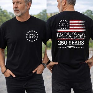May include: A black t-shirt featuring a white circular emblem with the text 1776 surrounded by stars on the front. The back displays a distressed American flag graphic with the text We The People, 250 YEARS, and 2026 in white lettering.