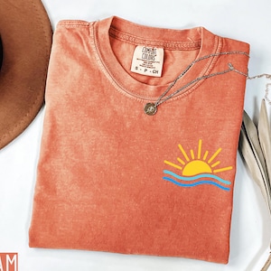 May include: Coral-colored Comfort Colors t-shirt with a small graphic of a yellow sun and blue waves. A silver necklace with a pendant rests on the shirt. The shirt has a small tag that reads "Comfort Colors".