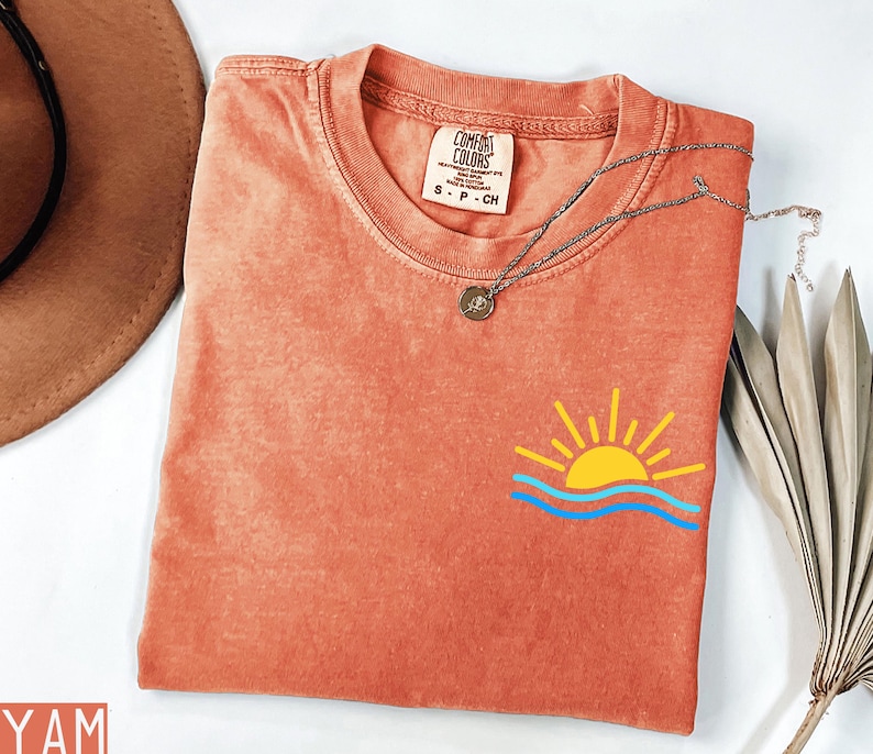May include: Coral-colored Comfort Colors t-shirt with a small graphic of a yellow sun and blue waves. A silver necklace with a pendant rests on the shirt. The shirt has a small tag that reads "Comfort Colors".