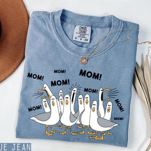 K&ouml;nnte beinhalten: Ein blaues Jeans-T-Shirt mit dem Wort "MOM!" mehrfach aufgedruckt. Das Shirt zeigt eine Grafik von Cartoon-M&ouml;wen mit ge&ouml;ffneten Schn&auml;beln. Eine goldene Halskette ist &uuml;ber dem Shirt drapiert.