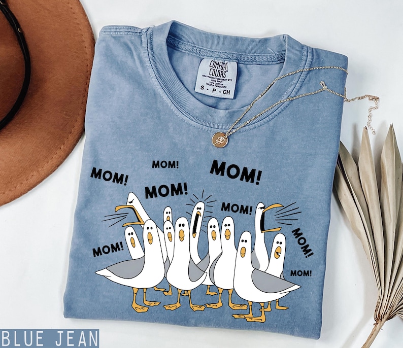 K&ouml;nnte beinhalten: Ein blaues Jeans-T-Shirt mit dem Wort "MOM!" mehrfach aufgedruckt. Das Shirt zeigt eine Grafik von Cartoon-M&ouml;wen mit ge&ouml;ffneten Schn&auml;beln. Eine goldene Halskette ist &uuml;ber dem Shirt drapiert.