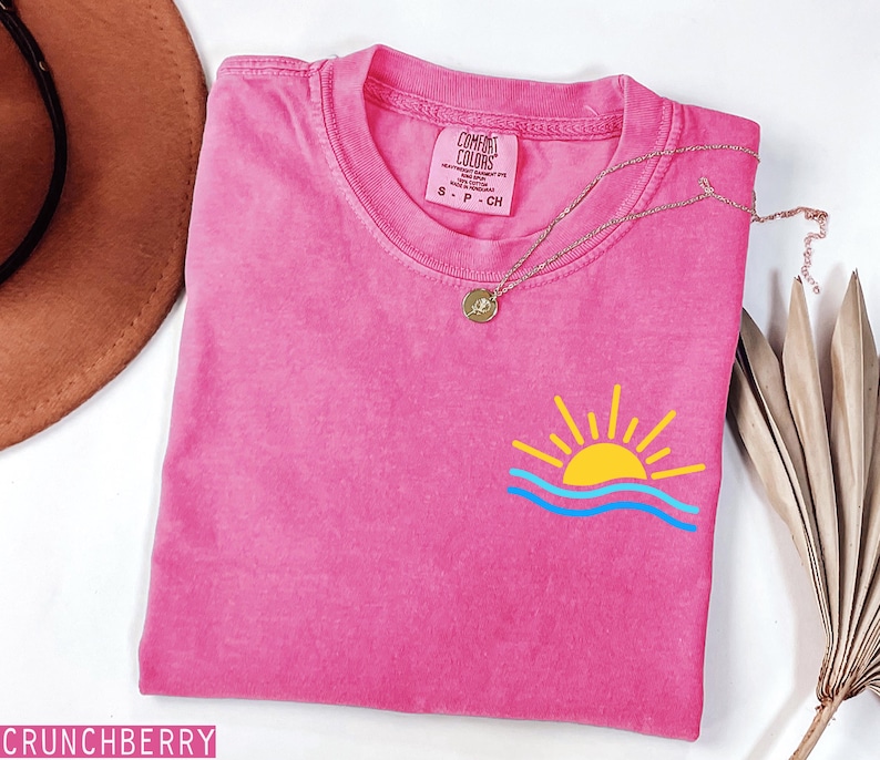 May include: A hot pink t-shirt with a small graphic of a yellow sun and blue waves. The shirt has a crew neck and short sleeves. A gold necklace with a pendant is draped across the shirt. The brand tag reads "Comfort Colors".
