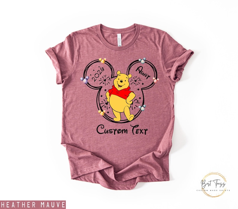 May include: Heather mauve t-shirt featuring a Winnie the Pooh design. The design includes a Mickey Mouse silhouette with the text "2026 Aunt" and "Custom Text". Winnie the Pooh is wearing a red shirt. The shirt has a crew neck.