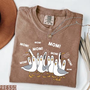 K&ouml;nnte beinhalten: Espressofarbenes T-Shirt mit einer Grafik von Zeichentrickm&ouml;wen, die "MOM!" in wei&szlig;em Text schreien. Das Shirt hat einen Rundhalsausschnitt und kurze &Auml;rmel. Eine silberne Halskette mit einem kleinen Anh&auml;nger liegt &uuml;ber dem Shirt.