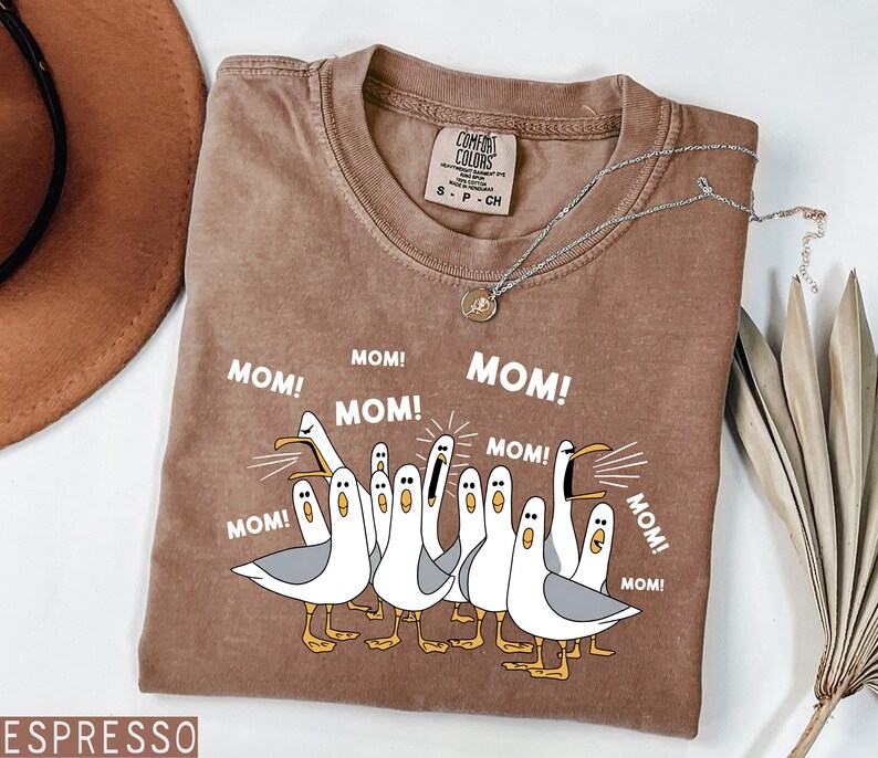 K&ouml;nnte beinhalten: Espressofarbenes T-Shirt mit einer Grafik von Zeichentrickm&ouml;wen, die "MOM!" in wei&szlig;em Text schreien. Das Shirt hat einen Rundhalsausschnitt und kurze &Auml;rmel. Eine silberne Halskette mit einem kleinen Anh&auml;nger liegt &uuml;ber dem Shirt.