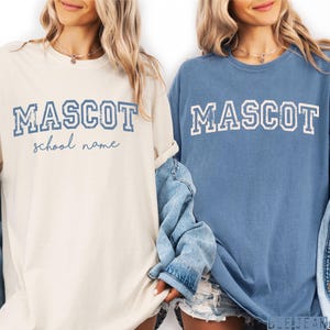 Puede incluir: Dos camisetas de manga corta y cuello redondo en color marfil y azul. La camiseta marfil presenta el texto azul MASCOT y school name en cursiva. La camiseta azul presenta el texto blanco MASCOT en una fuente universitaria desgastada.