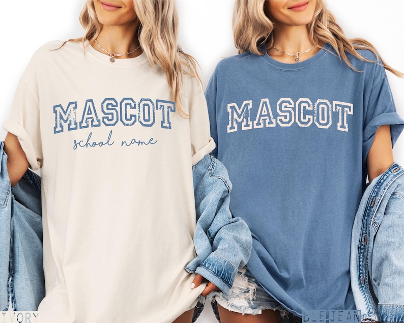 Puede incluir: Dos camisetas de manga corta y cuello redondo en color marfil y azul. La camiseta marfil presenta el texto azul MASCOT y school name en cursiva. La camiseta azul presenta el texto blanco MASCOT en una fuente universitaria desgastada.