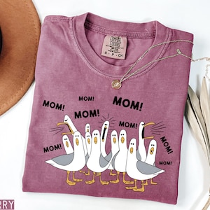 K&ouml;nnte beinhalten: Ein beerenfarbenes T-Shirt mit Rundhalsausschnitt und einer Grafik von Cartoon-M&ouml;wen, die "MOM!" in schwarzem Text schreien. Das Shirt hat eine lockere Passform und ein Comfort Colors-Etikett. Eine goldene Halskette mit Anh&auml;nger liegt auf dem Shirt.