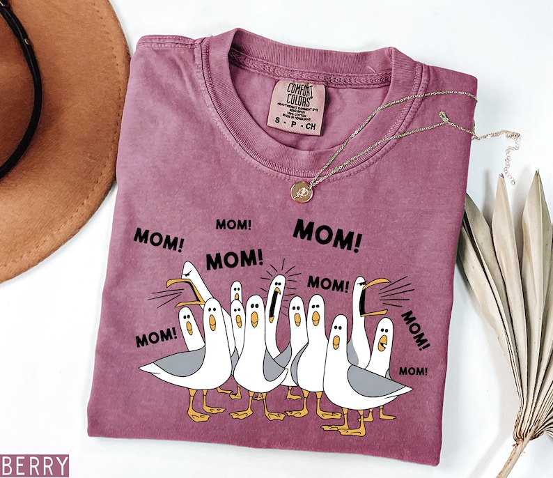 K&ouml;nnte beinhalten: Ein beerenfarbenes T-Shirt mit Rundhalsausschnitt und einer Grafik von Cartoon-M&ouml;wen, die "MOM!" in schwarzem Text schreien. Das Shirt hat eine lockere Passform und ein Comfort Colors-Etikett. Eine goldene Halskette mit Anh&auml;nger liegt auf dem Shirt.