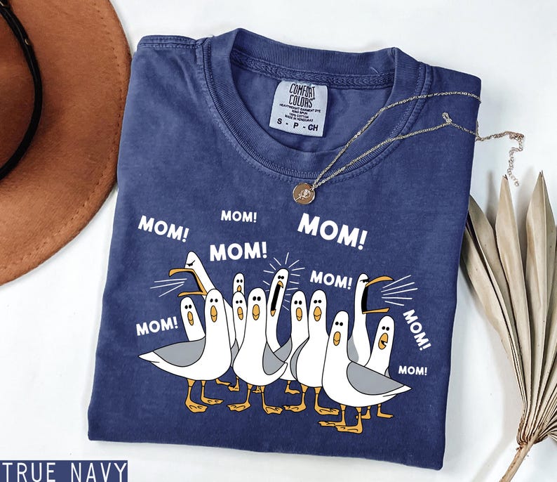 K&ouml;nnte beinhalten: Marineblaues T-Shirt mit einer Grafik von Cartoon-M&ouml;wen, die "MOM!" in wei&szlig; schreien. Das Shirt hat ein Comfort Colors-Etikett und wird mit einer goldenen Halskette kombiniert. Ein l&auml;ssiges Kleidungsst&uuml;ck.