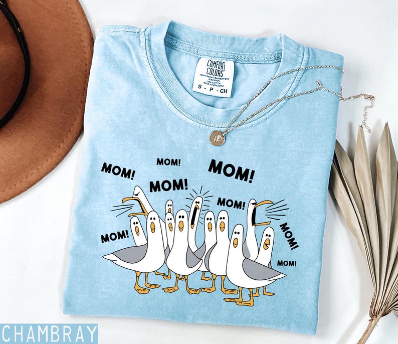 May include: Light blue Comfort Colors t-shirt with a graphic of seagulls yelling "MOM!". The shirt has a crew neck and short sleeves. A silver necklace with a pendant is draped on the shirt.