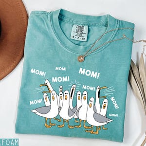 K&ouml;nnte beinhalten: T&uuml;rkisfarbenes T-Shirt mit einer Grafik von Cartoon-M&ouml;wen, die "MOM!" schreien. Das Shirt hat einen Rundhalsausschnitt und kurze &Auml;rmel. Eine goldene Halskette mit Anh&auml;nger befindet sich auf dem Shirt.