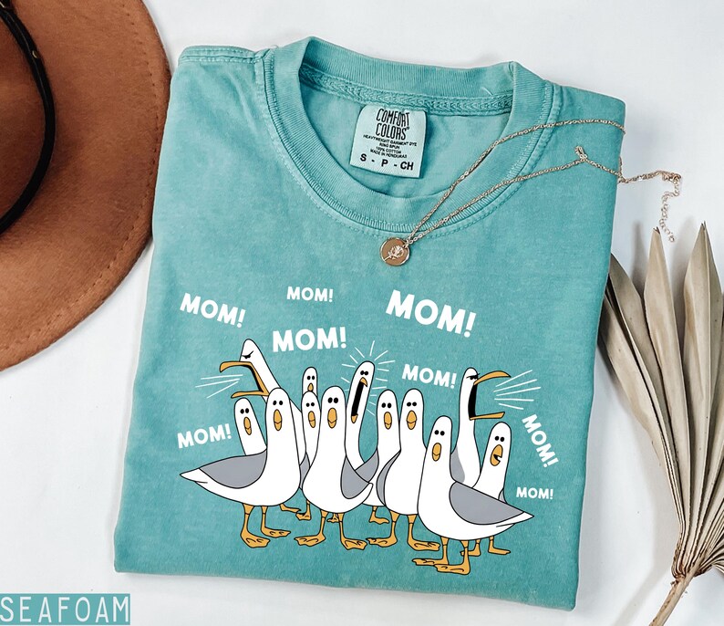 K&ouml;nnte beinhalten: T&uuml;rkisfarbenes T-Shirt mit einer Grafik von Cartoon-M&ouml;wen, die "MOM!" schreien. Das Shirt hat einen Rundhalsausschnitt und kurze &Auml;rmel. Eine goldene Halskette mit Anh&auml;nger befindet sich auf dem Shirt.