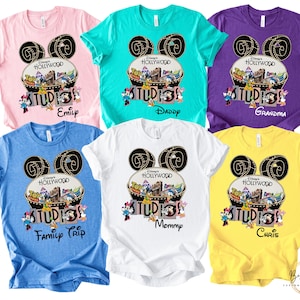 May include: A collection of colorful t-shirts featuring a Disney's Hollywood Studios design. Each shirt has a unique name printed below the graphic, including "Emilp", "Daddy", "Grandma", "Family Trip", "Mommy", and "Chris". The shirts are in various colors, including pink, teal, purple, blue, white, and yellow.