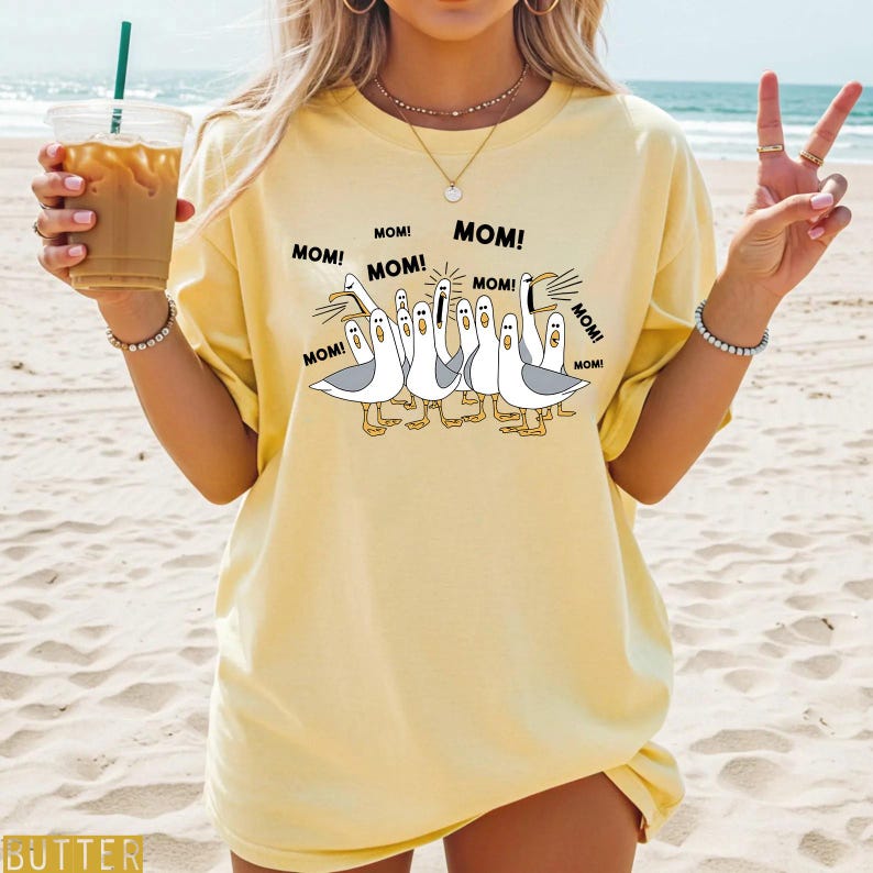 K&ouml;nnte beinhalten: Hellgelbes T-Shirt mit einem Cartoon-Design von M&ouml;wen, jeweils mit dem Wort "MOM!" dar&uuml;ber. Die M&ouml;wen sind wei&szlig; mit grauen Fl&uuml;geln und gelben F&uuml;&szlig;en. Das T-Shirt hat eine hellgelbe Farbe.