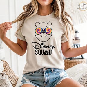 Puede incluir: Camiseta beige con una ilustraci&oacute;n de dibujos animados de Winnie the Pooh con gafas de sol te&ntilde;idas. Debajo, el texto "Disney Squad" con una silueta de castillo. La camiseta es de manga corta.