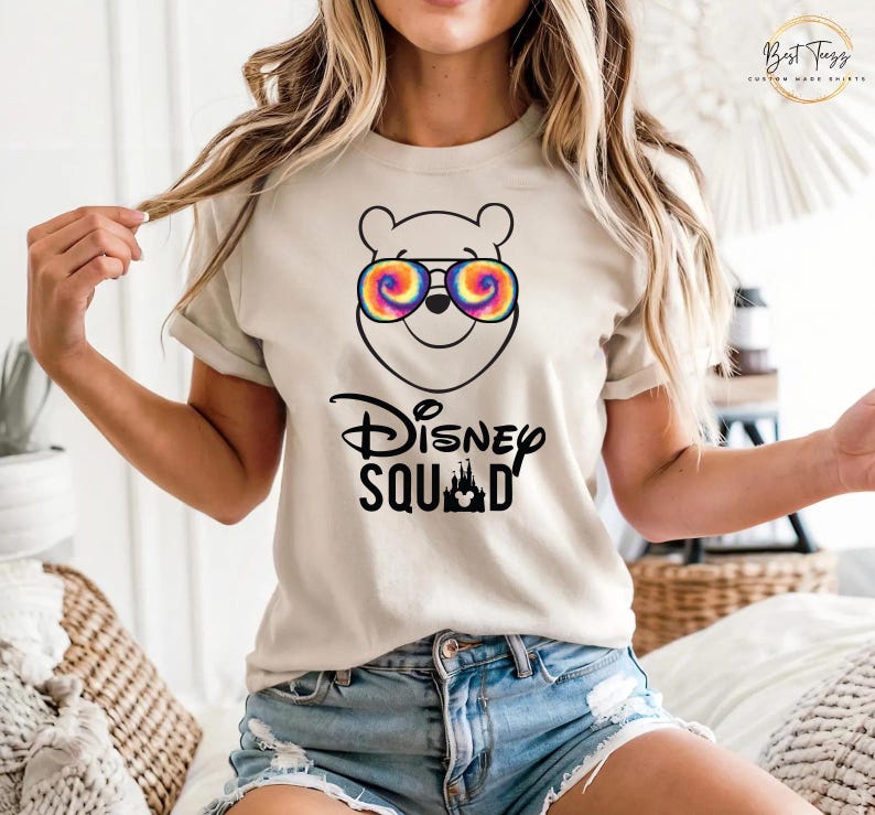 Puede incluir: Camiseta beige con una ilustraci&oacute;n de dibujos animados de Winnie the Pooh con gafas de sol te&ntilde;idas. Debajo, el texto "Disney Squad" con una silueta de castillo. La camiseta es de manga corta.