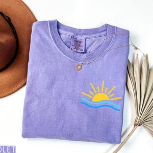 May include: A violet Comfort Colors t-shirt with a graphic of a yellow sun over blue waves. A gold necklace with a pendant rests on the shirt. A brown hat and dried palm leaf are in the background.