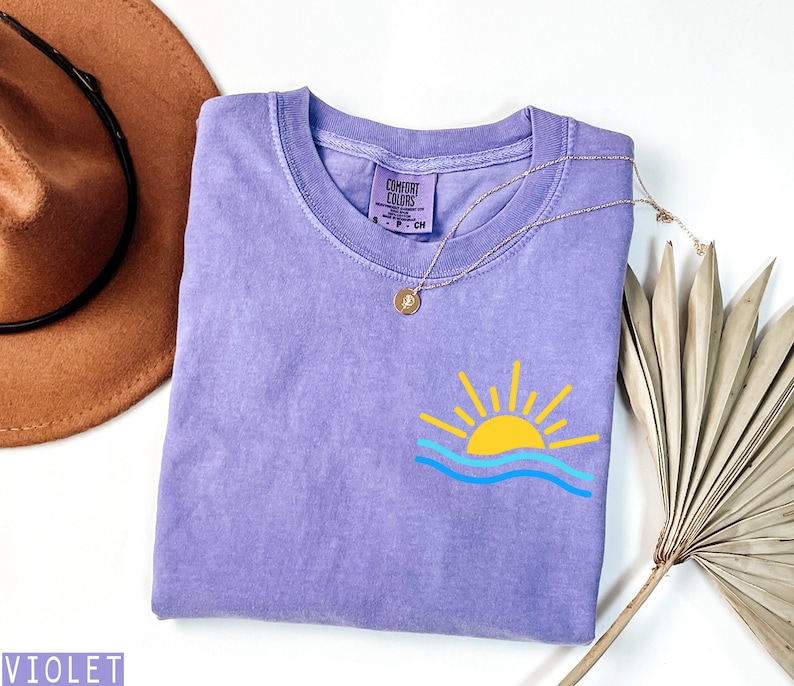 May include: A violet Comfort Colors t-shirt with a graphic of a yellow sun over blue waves. A gold necklace with a pendant rests on the shirt. A brown hat and dried palm leaf are in the background.