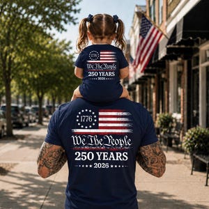 May include: A navy blue t-shirt featuring a graphic of the American flag, the number 1776, and the text We The People 250 YEARS 2026. The design is printed on the back of the shirt, showcasing a patriotic theme with white and red accents.