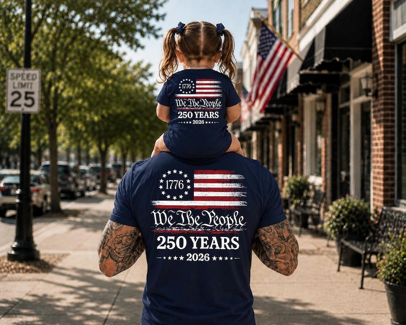 May include: A navy blue t-shirt featuring a graphic of the American flag, the number 1776, and the text We The People 250 YEARS 2026. The design is printed on the back of the shirt, showcasing a patriotic theme with white and red accents.
