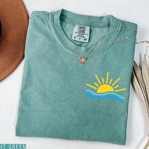 May include: Light green t-shirt with a yellow sun and blue wave graphic. The shirt has a crew neck and short sleeves. A silver necklace with a small pendant is draped across the shirt.