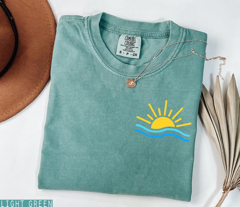 May include: Light green t-shirt with a yellow sun and blue wave graphic. The shirt has a crew neck and short sleeves. A silver necklace with a small pendant is draped across the shirt.