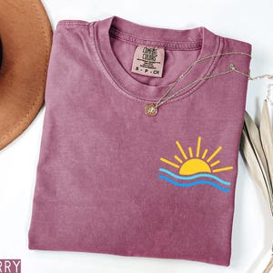 May include: Berry-colored t-shirt with a crew neck and short sleeves. The shirt features a small graphic of a yellow sun with blue waves. A gold necklace with a pendant is draped across the shirt. The shirt is labeled "Comfort Colors".