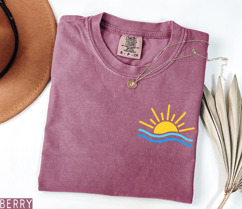 May include: Berry-colored t-shirt with a crew neck and short sleeves. The shirt features a small graphic of a yellow sun with blue waves. A gold necklace with a pendant is draped across the shirt. The shirt is labeled "Comfort Colors".
