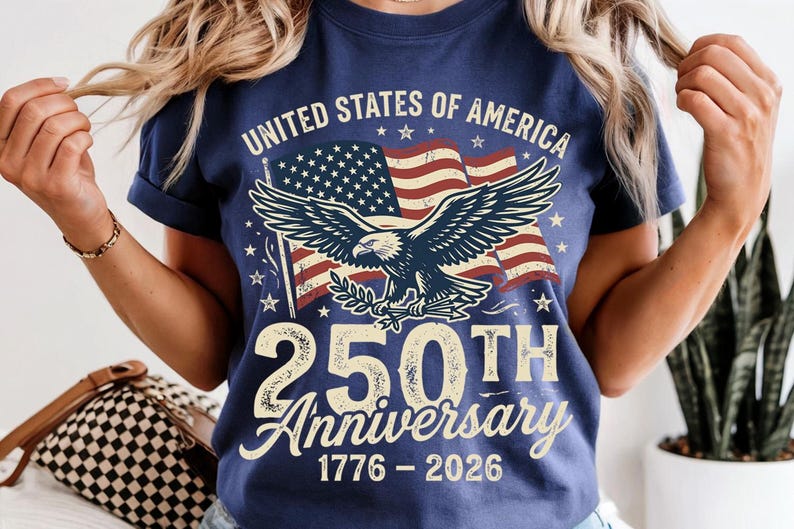 Pode incluir: Uma t-shirt azul-marinho com um gr&aacute;fico de uma &aacute;guia-de-cabe&ccedil;a-branca em frente a uma bandeira americana. O texto diz UNITED STATES OF AMERICA, 250TH Anniversary e 1776 - 2026. O design usa uma fonte de estilo vintage e cores patri&oacute;ticas.