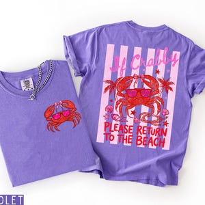 K&ouml;nnte beinhalten: Zwei violette T-Shirts mit Krabben-Grafiken. Ein Shirt hat eine kleine Krabben-Grafik auf der Vorderseite, das andere ein gr&ouml;&szlig;eres Krabben-Design auf dem R&uuml;cken mit dem Text "If Crabby Please Return to the Beach". Eine silberne Halskette befindet sich auf einem Shirt.