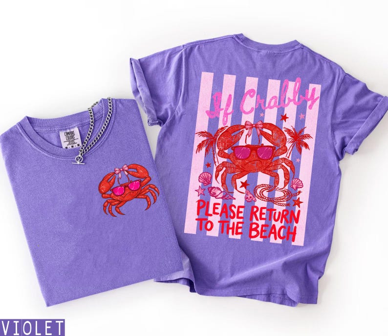 K&ouml;nnte beinhalten: Zwei violette T-Shirts mit Krabben-Grafiken. Ein Shirt hat eine kleine Krabben-Grafik auf der Vorderseite, das andere ein gr&ouml;&szlig;eres Krabben-Design auf dem R&uuml;cken mit dem Text "If Crabby Please Return to the Beach". Eine silberne Halskette befindet sich auf einem Shirt.