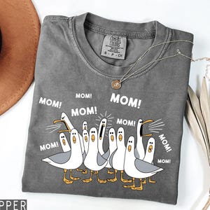K&ouml;nnte beinhalten: Graues Comfort Colors T-Shirt mit einem Cartoon von M&ouml;wen und dem Wort "MOM!" dar&uuml;ber. Die M&ouml;wen sind wei&szlig; mit grauen Fl&uuml;geln und gelben F&uuml;&szlig;en. Eine silberne Halskette mit Anh&auml;nger befindet sich auf dem T-Shirt.