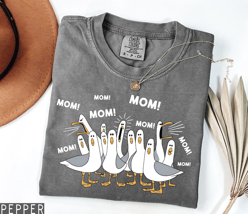 K&ouml;nnte beinhalten: Graues Comfort Colors T-Shirt mit einem Cartoon von M&ouml;wen und dem Wort "MOM!" dar&uuml;ber. Die M&ouml;wen sind wei&szlig; mit grauen Fl&uuml;geln und gelben F&uuml;&szlig;en. Eine silberne Halskette mit Anh&auml;nger befindet sich auf dem T-Shirt.