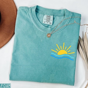 May include: Seafoam green t-shirt with a yellow sun and blue wave graphic. The shirt has a crew neck and short sleeves. A gold necklace with a pendant is draped across the shirt. A brown hat and dried palm leaf are in the background.