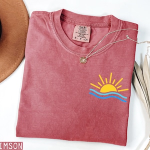 May include: Crimson-colored t-shirt with a graphic of a yellow sun and blue waves. The shirt has a crew neck and a small tag near the neckline. A silver necklace with a round pendant is draped across the shirt.
