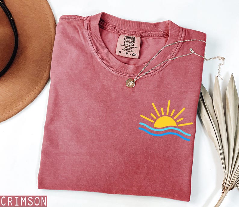 May include: Crimson-colored t-shirt with a graphic of a yellow sun and blue waves. The shirt has a crew neck and a small tag near the neckline. A silver necklace with a round pendant is draped across the shirt.