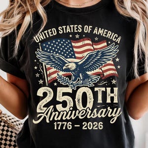 Pode incluir: Uma t-shirt preta de manga curta com um gr&aacute;fico de uma &aacute;guia-de-cabe&ccedil;a-branca em frente a uma bandeira dos Estados Unidos. O texto diz UNITED STATES OF AMERICA, 250TH Anniversary e 1776-2026. O design apresenta uma estampa desgastada vintage.