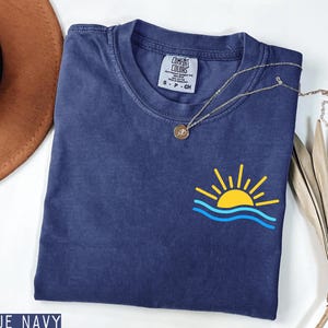 May include: A navy blue t-shirt with a yellow and blue sun and wave graphic. The shirt has a small tag that says "Comfort Colors". A gold necklace with a pendant is draped across the shirt. The words "True Navy" are at the bottom.