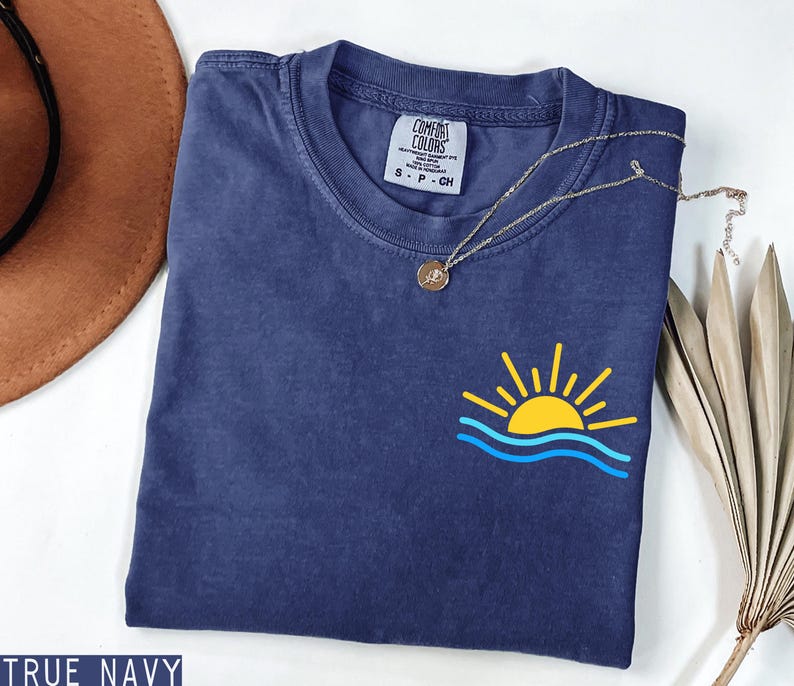 May include: A navy blue t-shirt with a yellow and blue sun and wave graphic. The shirt has a small tag that says "Comfort Colors". A gold necklace with a pendant is draped across the shirt. The words "True Navy" are at the bottom.