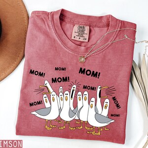 K&ouml;nnte beinhalten: Ein weinrotes T-Shirt mit einer Grafik von Cartoon-M&ouml;wen und dem Wort "MOM!" wiederholt. Das T-Shirt hat einen Rundhalsausschnitt und kurze &Auml;rmel. Eine goldene Halskette mit Anh&auml;nger ist auf dem T-Shirt drapiert.