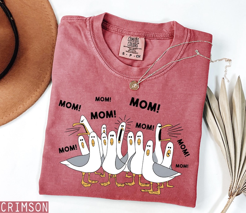 K&ouml;nnte beinhalten: Ein weinrotes T-Shirt mit einer Grafik von Cartoon-M&ouml;wen und dem Wort "MOM!" wiederholt. Das T-Shirt hat einen Rundhalsausschnitt und kurze &Auml;rmel. Eine goldene Halskette mit Anh&auml;nger ist auf dem T-Shirt drapiert.